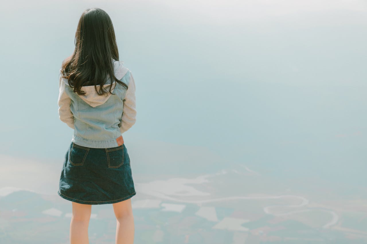 Back view of a young girl in a denim jacket gazing at a vast scenic landscape, capturing a sense of freedom and adventure.